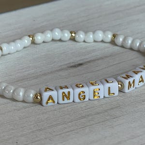 May include: White beaded bracelet with gold lettering spelling "Angel Mama".