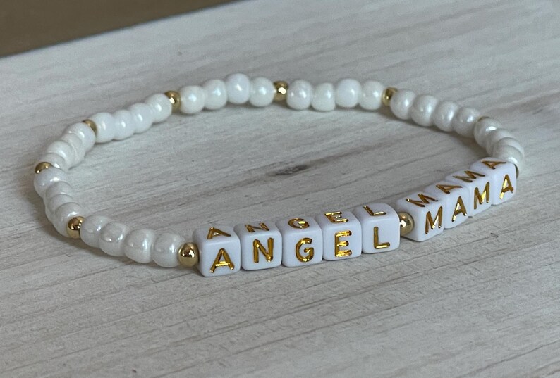 May include: White beaded bracelet with gold lettering spelling "Angel Mama".