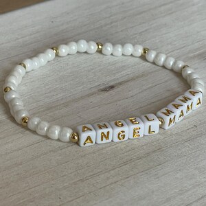 May include: White beaded bracelet with gold accents and white letter beads spelling "ANGEL MAMA".