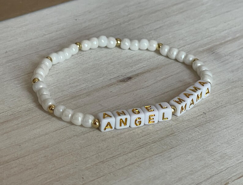 May include: White beaded bracelet with gold accents and white letter beads spelling "ANGEL MAMA".
