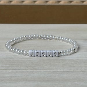 May include: A stretch bracelet featuring clear, translucent beads and five white square beads in the center. Each white bead displays a single black letter, collectively spelling the word JESUS. The design includes small silver-toned metallic spacers.