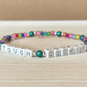 May include: A beaded bracelet with the words "TOUGH COOKIE" spelled out in white square beads. The bracelet features colorful beads in various colors, including pink, blue, green, and yellow, and is strung on an elastic band.