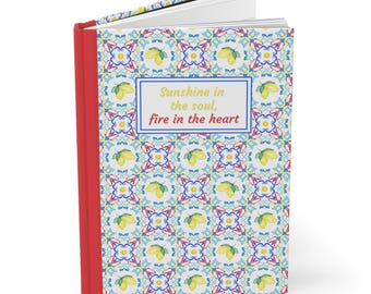 May include: Open journal with a red spine and a patterned cover. The cover features a repeating design of lemons and blue and yellow floral patterns. The text "Sunshine in the soul, fire in the heart" is displayed on the cover.