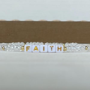 May include: A beaded bracelet featuring clear, translucent beads interspersed with small gold-toned metallic beads. The center of the bracelet displays five white square beads with the gold-colored letters F, A, I, T, and H printed on them.