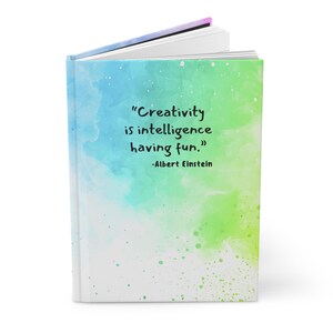 May include: A white journal with a watercolor background of blue, green, and white. The quote "Creativity is intelligence having fun." by Albert Einstein is printed on the front cover.