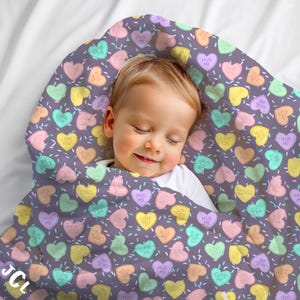 May include: A baby wrapped in a gray blanket patterned with colorful candy hearts. The hearts have phrases like "Love You" and "Hug Me." The blanket is on a white surface. The baby is sleeping.