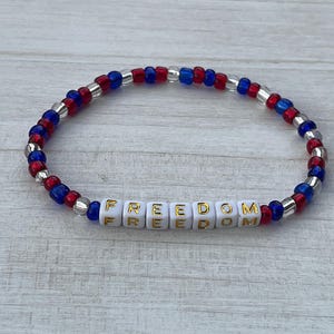 Puede incluir: Una pulsera de cuentas rojas, blancas y azules con la palabra "FREEDOM" escrita en letras doradas sobre cuentas blancas.