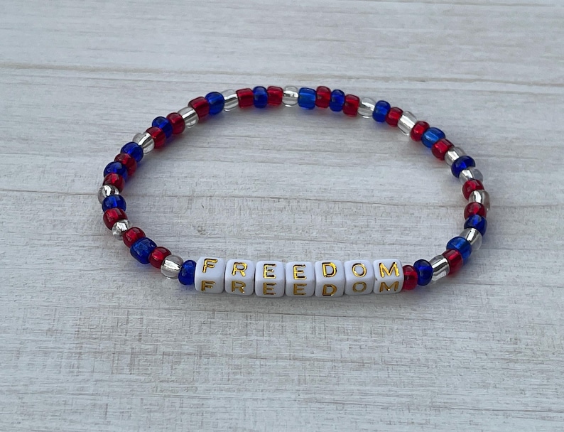 Puede incluir: Una pulsera de cuentas rojas, blancas y azules con la palabra "FREEDOM" escrita en letras doradas sobre cuentas blancas.