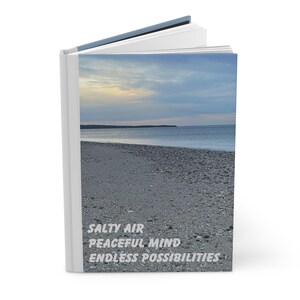 May include: A book with a white cover and a blue spine. The cover features a photo of a beach with a cloudy sky and the text "Salty Air Peaceful Mind Endless Possibilities".