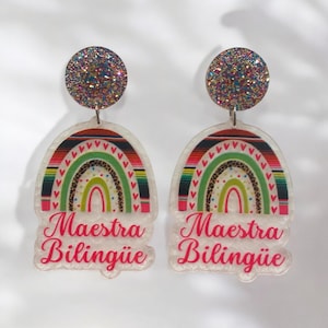 May include: A pair of rainbow-themed earrings with the text "Maestra Bilingüe" in pink. The earrings feature a rainbow with a colorful Mexican-style pattern and glitter accents.