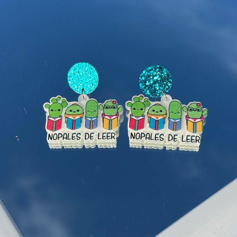 Op de afbeelding: Een paar hangende oorbellen met vier groene cactusfiguren die kleurrijke boeken vasthouden. De oorbellen hebben een turquoise glitteraccent en de tekst "Nopales de Leer" onderaan.