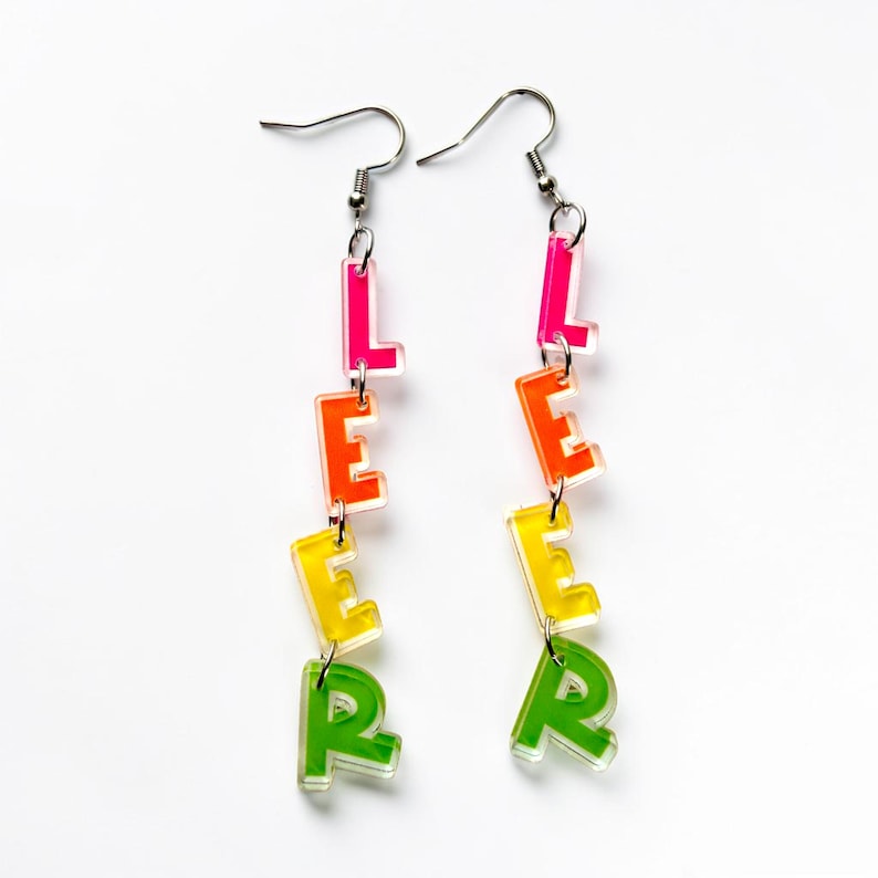 May include: A pair of dangling earrings spelling out the word "LEER" in a gradient of colors. The letters are in a clear, blocky font, with each letter a different color: pink, orange, yellow, and green. The earrings have silver-colored hooks.