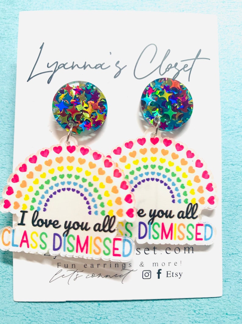 Op de afbeelding: Regenboogvormige oorringen in de vorm van een hart met de tekst "I love you all CLASS DISMISSED" en "e you all DISMISSED" op elke oorring. De oorringen zijn gemaakt van helder acryl met glittersterren aan de bovenkant.