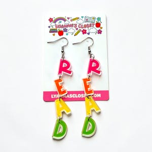 May include: Colorful dangle earrings spelling out the word "READ". The letters are in bright pink, orange, yellow, and green. The earrings hang from silver hooks and are displayed on a white card with the text "LYANNA'S CLOSET".