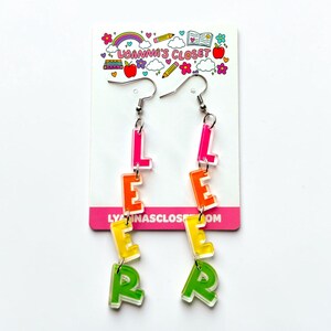 May include: A pair of dangle earrings spelling out the word "LEARN" in colorful, transparent acrylic letters. The letters are in shades of pink, orange, yellow, and green, suspended from silver-tone ear wires. The earrings are displayed on a white card with a colorful design.