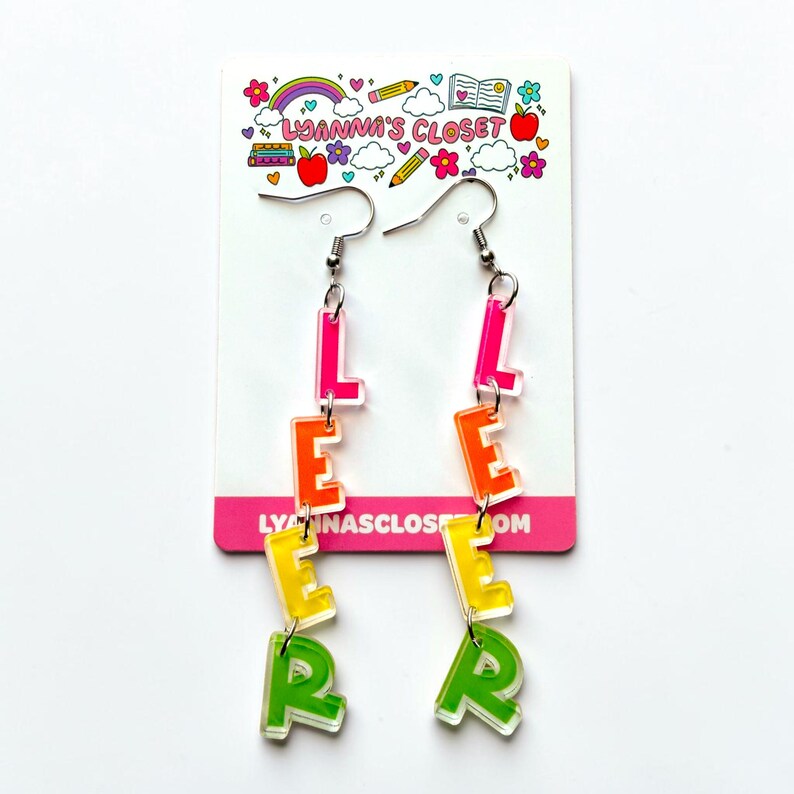 May include: A pair of dangle earrings spelling out the word "LEARN" in colorful, transparent acrylic letters. The letters are in shades of pink, orange, yellow, and green, suspended from silver-tone ear wires. The earrings are displayed on a white card with a colorful design.