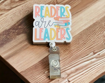 Puede incluir: Un portatarjetas blanco con el texto "READERS are LEADERS" en letras coloridas. El diseño incluye una pila de libros. El portatarjetas tiene un soporte de identificación de plástico transparente y un clip de metal.