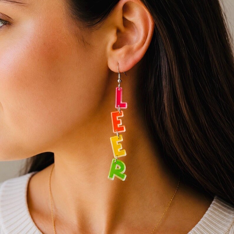 May include: A pair of dangling earrings spelling out the word "LEER" in colorful, stacked letters. The letters are in shades of pink, orange, yellow, and green, with a clear outline. The earrings are suspended from silver hooks.