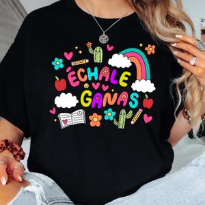 May include: Black t-shirt with a colorful graphic design featuring the text "Échale Ganas" surrounded by rainbows, hearts, cacti, and other fun illustrations.