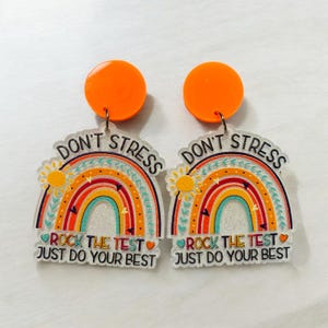 May include: A pair of clear acrylic earrings with a rainbow design and the text "Don't Stress, Rock the Test, Just Do Your Best". The earrings are attached to orange round discs.