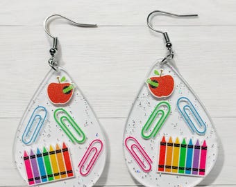 Può includere: Un paio di orecchini trasparenti a forma di lacrima con ganci argentati. Gli orecchini sono pieni di glitter e presentano ciondoli colorati a tema scolastico, tra cui una mela rossa, graffette e matite colorate.
