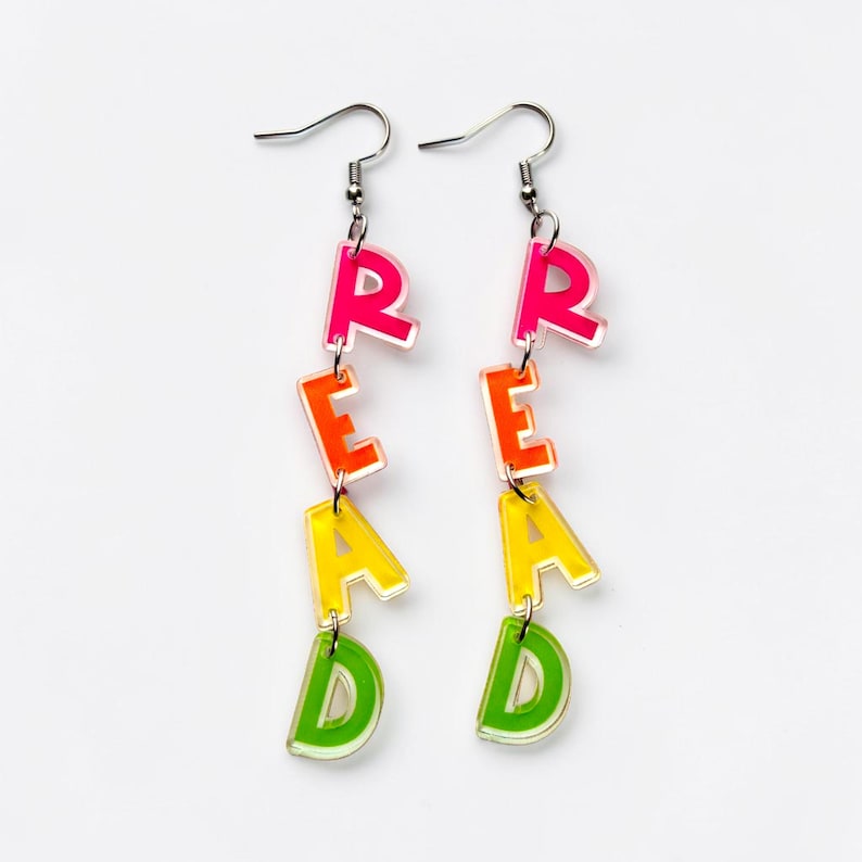 May include: A pair of dangle earrings spelling out the word "READ" in colorful, layered acrylic letters. The letters are in a gradient of pink, orange, yellow, and green, suspended from silver-tone ear wires.