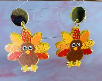 May include: A pair of gold-tone earrings featuring a cartoon turkey design. The turkeys are brown with orange, yellow, and red feathers.
