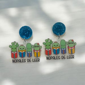 Op de afbeelding: Een paar blauwe glitter oorbellen met een transparante hanger met vijf cartooncactussen die een bril dragen en boeken vasthouden. De hanger heeft de tekst "Nopales de Leer".