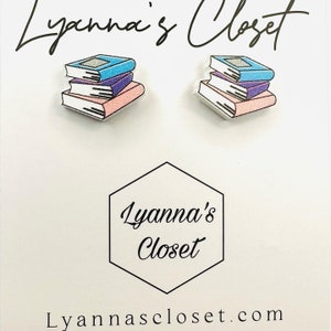 May include: A pair of book stack earrings. The earrings are made of wood and painted to look like a stack of books. The books are blue, purple, and pink. The earrings are a fun and whimsical way to show your love of reading.