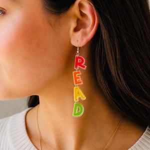 May include: Colorful dangle earrings spelling out the word "READ". The letters are in a gradient of red, orange, yellow, and green. The earrings are suspended from silver-colored hooks.