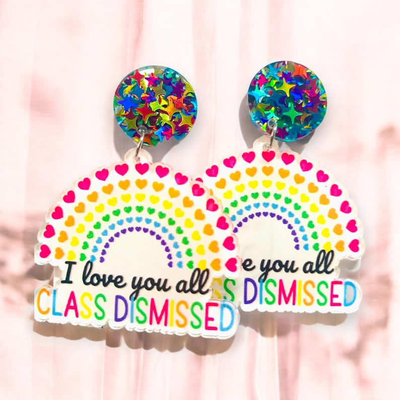 Op de afbeelding: Regenboog-oorbellen met een hart- en regenboogontwerp. De oorbellen bevatten de tekst "I love you all CLASS DISMISSED" en een glinsterende sterbedel.
