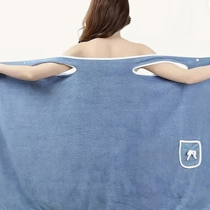 May include: A blue wrap-around towel with white trim and armholes. The towel has a small pocket with a white bow design. The person is holding the towel open, showcasing the design and fabric.