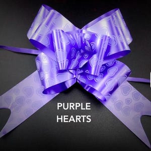 May include: A shiny purple gift bow featuring a repeating pattern of heart outlines. The ribbon has multiple loops and two long, notched tails extending downwards. The text PURPLE HEARTS is printed in white capital letters below the bow.