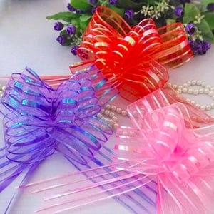 May include: Three decorative pull bows in vibrant translucent colors: one bright red, one deep purple, and one soft pink. Each bow features a multi-layered, looped design with long, thin ribbon tails extending from the base of the structure.