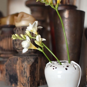 Op de afbeelding: Witte keramische vaas met een uniek ontwerp van gaten over het hele oppervlak. De vaas is gevuld met witte en gele freesia bloemen.