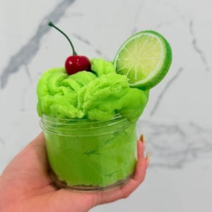 May include: A clear plastic jar filled with bright green textured slime. The top of the slime is decorated with a single red cherry with a green stem and a realistic slice of lime positioned upright on the side of the container.