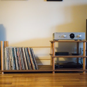 May include: A wooden record player stand with three shelves, a silver amplifier, and two black speakers. The stand is holding a collection of vinyl records.