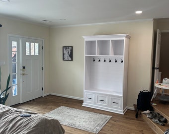 May include: A white entryway storage unit with a bench, hooks, and cubbies. The unit is against a cream-colored wall, with a rug on the wood floor. A front door is visible in the background.