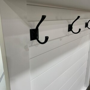 以下が含まれることがあります： 白い壁掛けコートラック、黒い金属製のフックが3つ付いています。フックは白い水平板張りの表面に取り付けられています。フックは湾曲したデザインです。