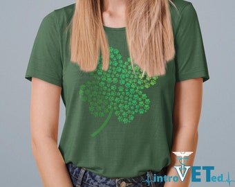 Può includere: Una maglietta verde con un design a trifoglio a quattro foglie fatto di impronte di zampe. Il trifoglio è in diverse tonalità di verde, con uno stelo. La maglietta è indossata da una persona con lunghi capelli biondi.