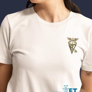 May include: White t-shirt featuring a gold and green veterinary symbol with a frog. The shirt also has the text "introVETed" in blue. The shirt is a crew neck and has short sleeves.