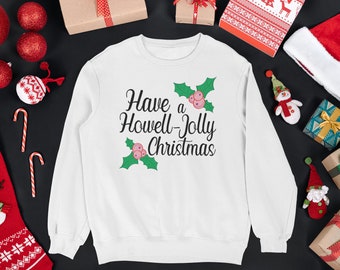 Può includere: Felpa bianca natalizia con foglie di agrifoglio verdi e bacche rosa. Il testo recita "Have a Howell-Jolly Christmas".