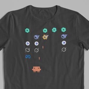 May include: Dark grey t-shirt with a pixelated video game design. The design features a brown spaceship, geometric shapes, and explosions in blue, yellow, and green. The shirt has a classic crew neck.