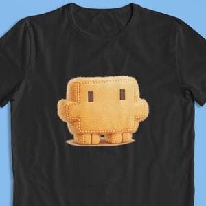 Puede incluir: Una camiseta negra de manga corta con un gráfico centrado de un personaje rectangular, difuso y de color naranja claro, con dos pequeños ojos cuadrados de color marrón oscuro, brazos cortos y dos piernas robustas de aspecto afelpado.