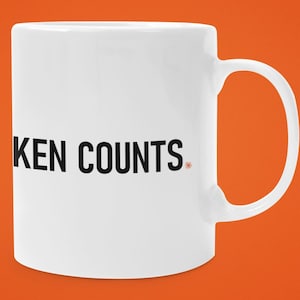 May include: A white ceramic mug with a handle on the right side. The text KEN COUNTS is printed in black capital letters on the front of the mug. There is a small orange circular symbol located to the right of the word COUNTS on the mug surface.