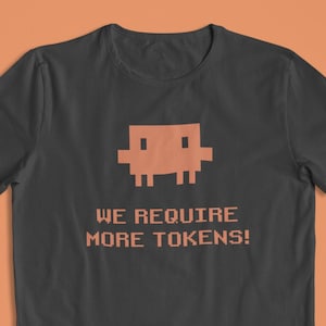 Puede incluir: Camiseta gris oscuro con un gráfico pixelado naranja y el texto "WE REQUIRE MORE TOKENS!". El diseño presenta una estética de videojuego retro, ideal para jugadores y fanáticos del arte digital vintage.