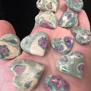 May include: A collection of heart-shaped stones in shades of green, beige, and pink. The stones have a speckled appearance, with the pink areas concentrated in the center of each heart. The stones are smooth and polished.