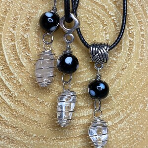 May include: A black leather cord necklace with a silver pendant featuring a clear quartz crystal encased in wire. The necklace also includes two dangling charms, each with a black and white glass bead and a clear quartz crystal encased in wire.