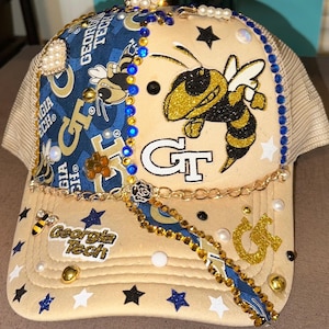 May include: Tan and navy blue trucker hat with a gold and black bee graphic, embellished with pearls, rhinestones, and gold accents. The hat features the text "Georgia Tech" and the letters "GT".