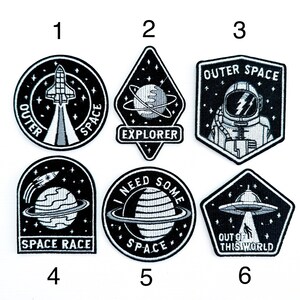 May include: A collection of six black and white embroidered space-themed patches. Designs include a rocket, planets, an astronaut, and the phrases "Outer Space," "Explorer," "Space Race," "I Need Some Space," and "Out of This World."
